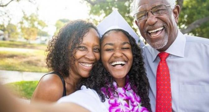 young female graduate in robes and gowns smiling and taking a selfie with parents